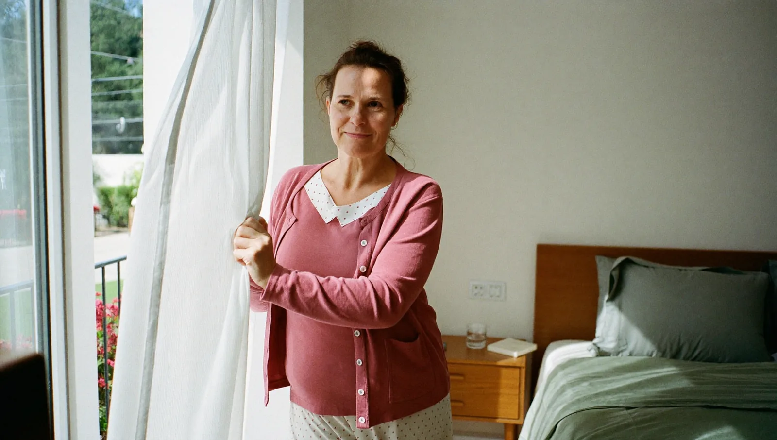 Žena kolem sedmačtyřiceti v pyžamu a svetru odhrnuje ráno závěs u okna světlé ložnice, za ní ustlaná postel